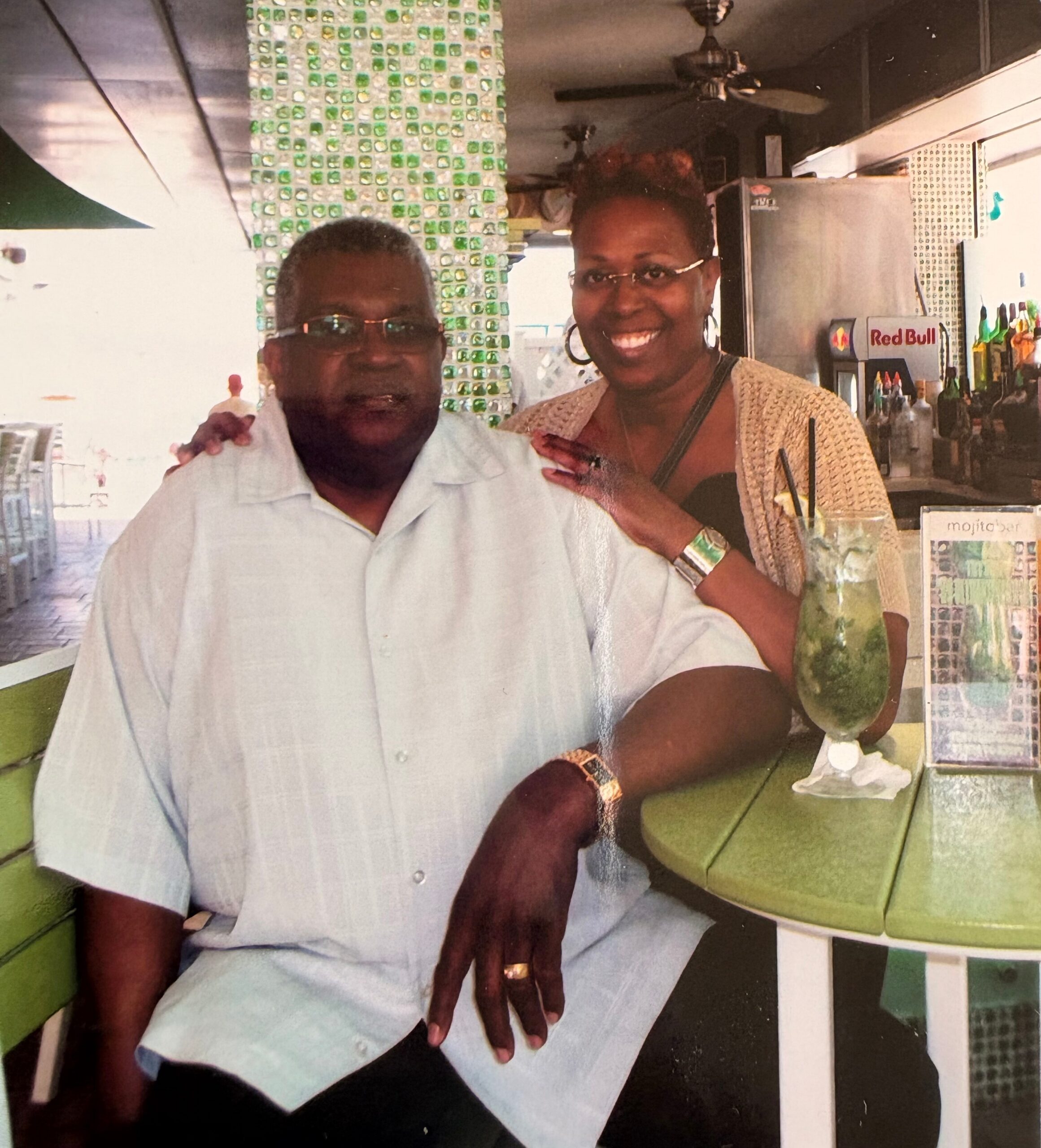 Smiling man in a light shirt and a smiling woman with short hair and glasses pose at a bar. A green cushioned seat and mosaic wall are behind them, with a drink on the counter.