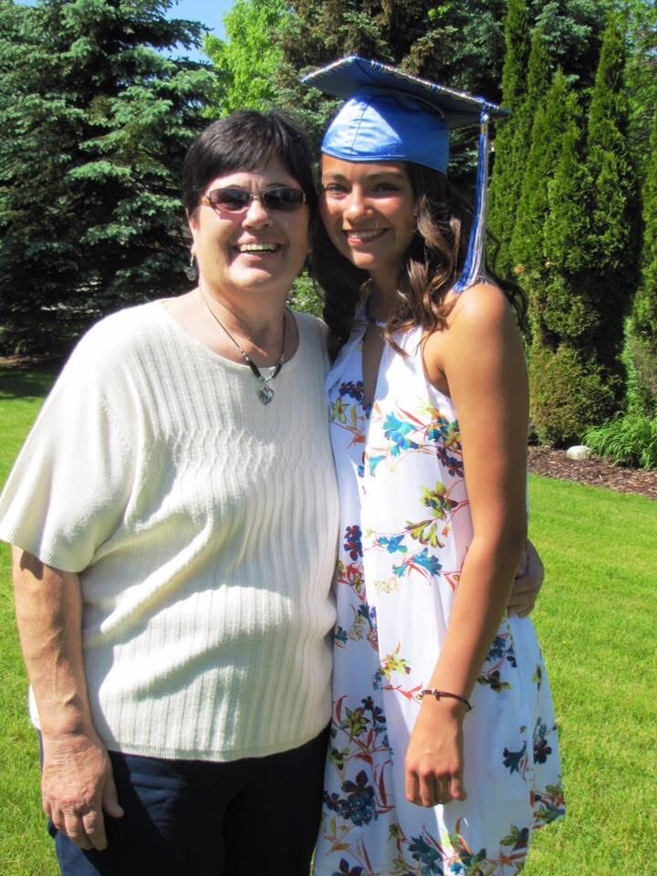 A graduate in a blue cap and floral dress with an older woman in a cream sweater outdoors, smiling together in a garden.