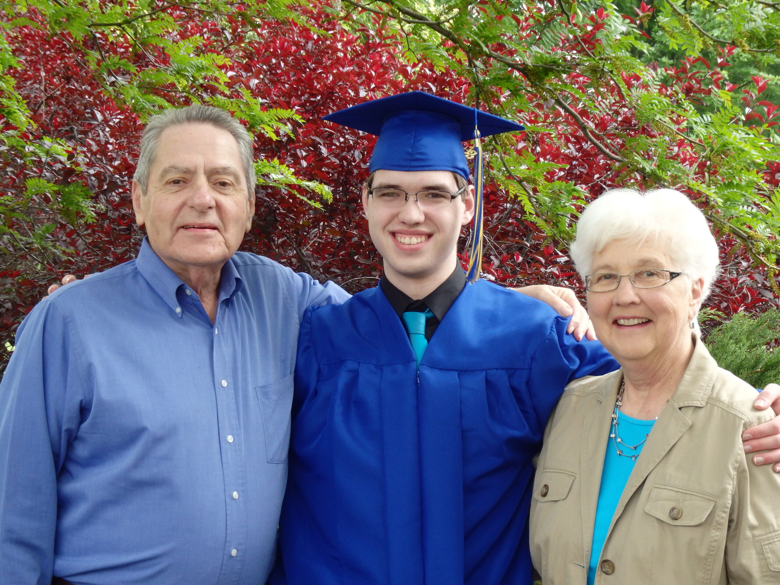 Graduate in royal blue cap and gown posing with two smiling adults outdoors among green and red foliage.