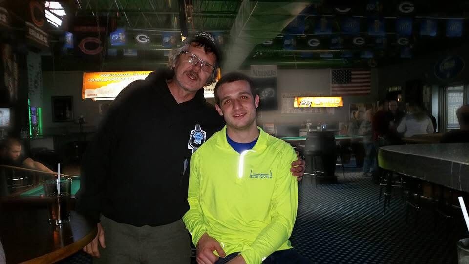 Two men pose together in a dimly lit billiards bar; one in a black hoodie and cap, the other in a neon yellow jacket, smiling at the camera.