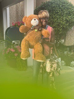Person sitting on a porch or patio, holding a large brown teddy bear with a white snout, with greenery framing the scene in the foreground.