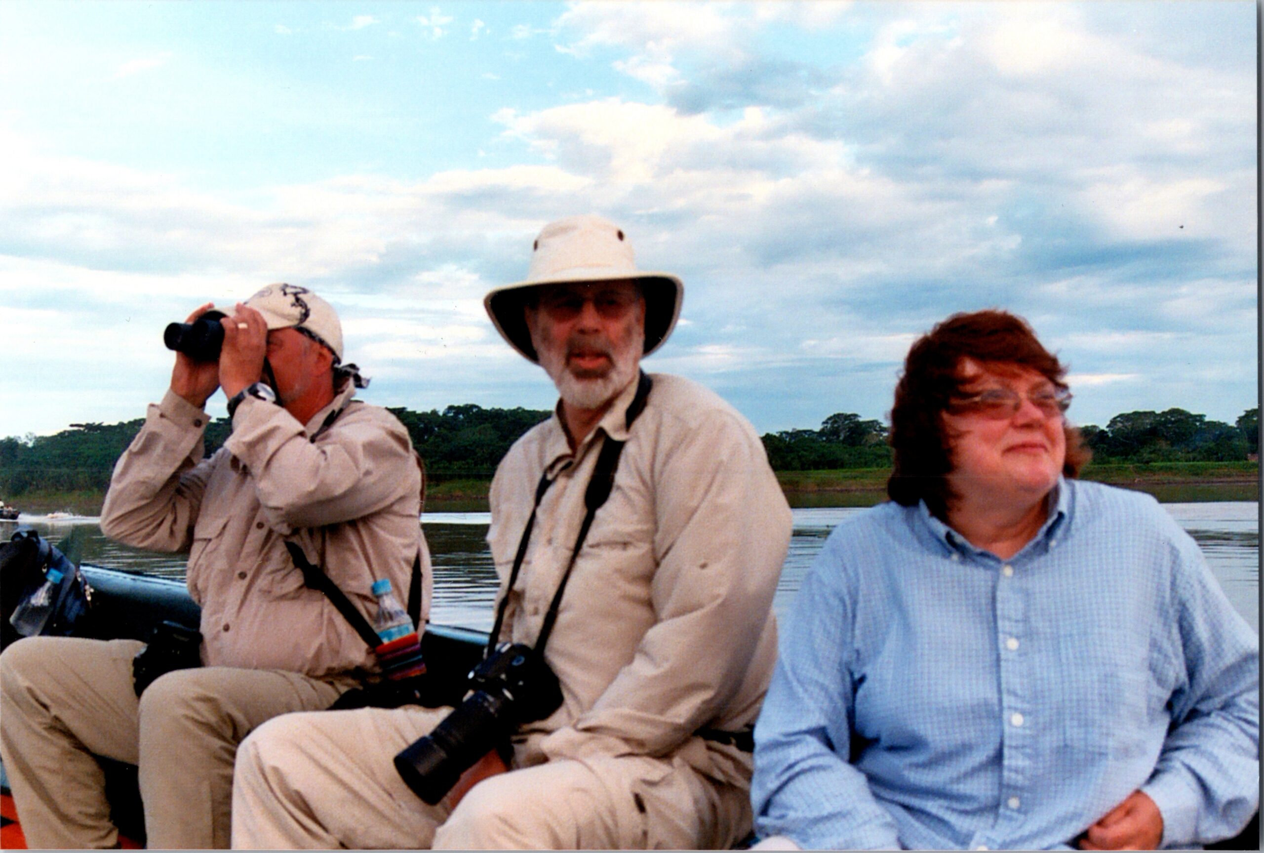 Three people in a boat on a lake; man on the left uses binoculars, the center man wears a wide-brimmed hat, and a woman in a blue shirt sits to the right.