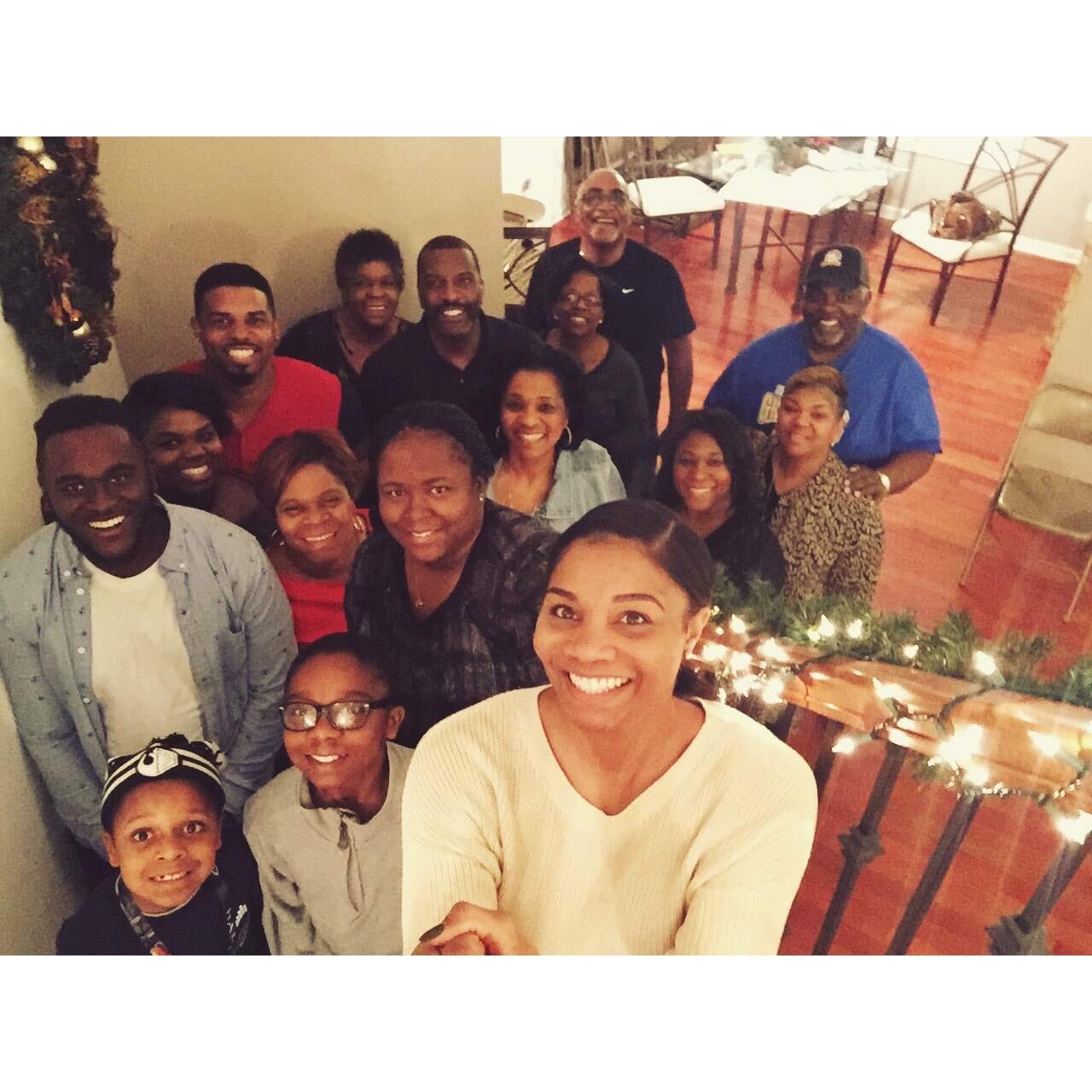 Smiling group of diverse friends and family posing for a selfie at a holiday gathering, Christmas lights along a railing in the foreground.