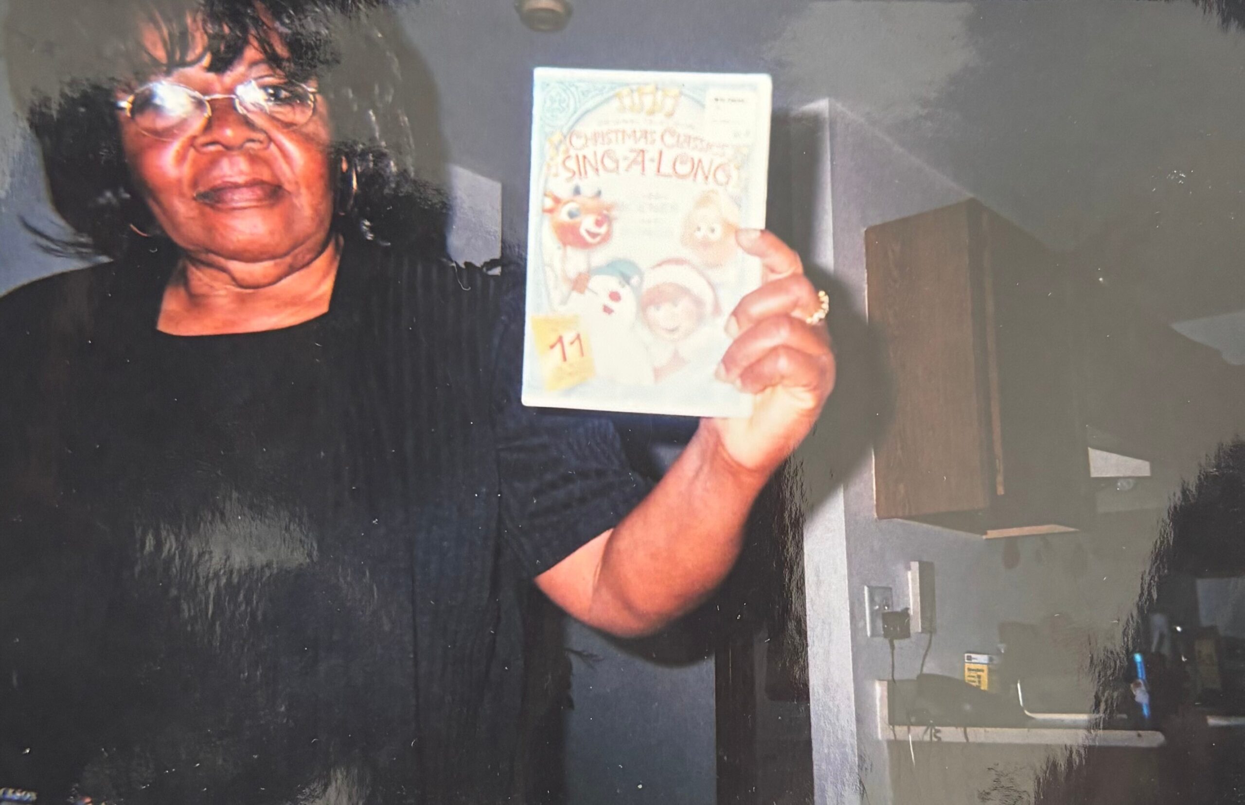 Older woman wearing glasses holds up a colorful Christmas Sing-Along book with cartoon characters in a doorway hallway.] ,