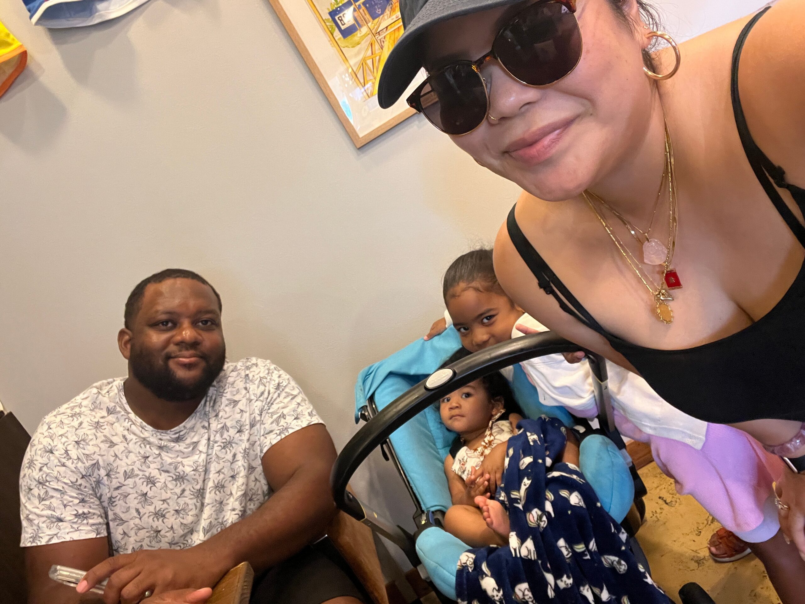 Person taking a selfie at a casual gathering; woman in foreground wearing sunglasses and gold jewelry smiles at the camera, others in a stroller nearby.