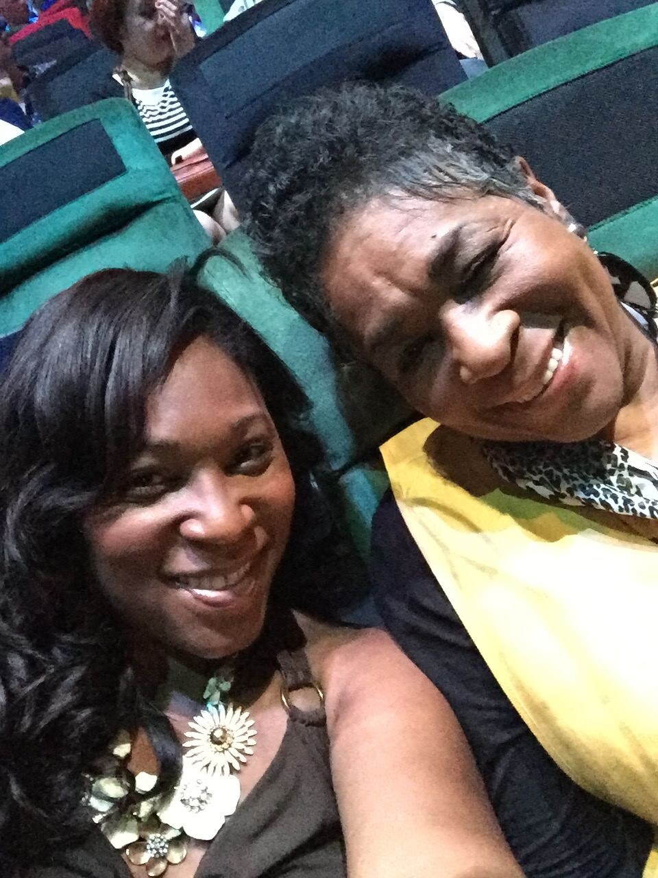 Two women sit close together in a theater, smiling for a selfie; one wears a chunky floral necklace and a brown top, the other a yellow scarf and patterned blouse.