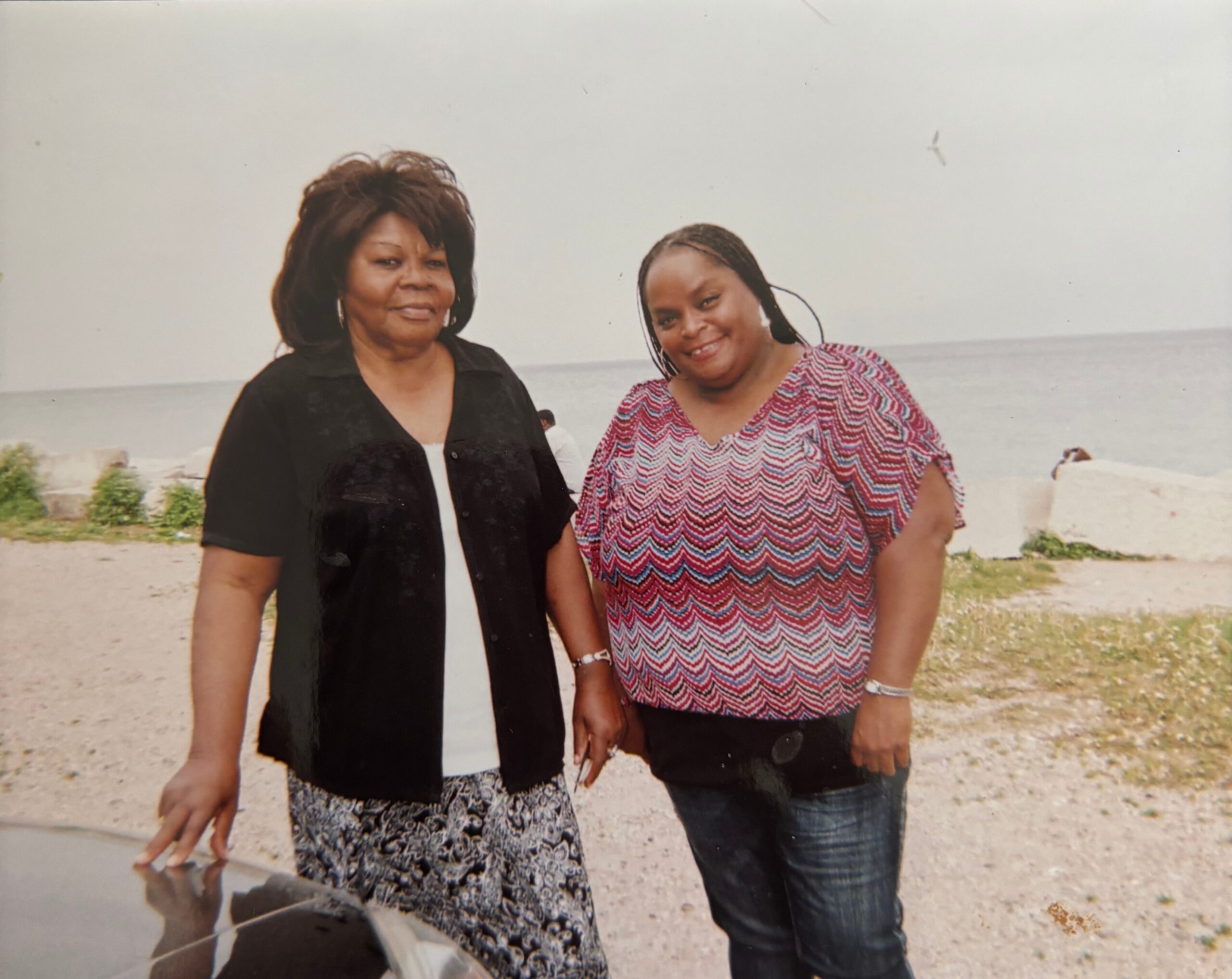 Two women standing on a beach, smiling at the camera with the ocean behind them.