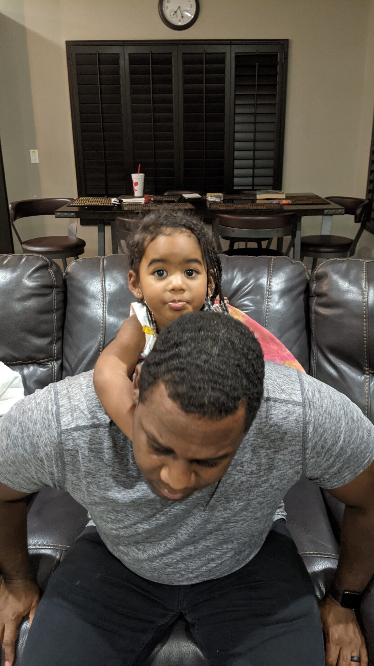 Young girl sits on a man's shoulders on a brown leather couch in a living room, looking at the camera.