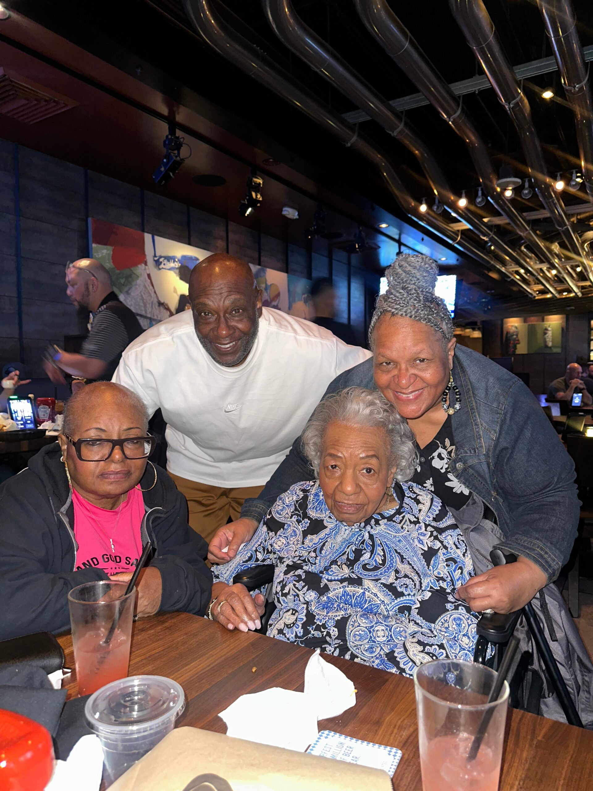 Group of four adults at a restaurant posing for a photo, smiling at the camera with drinks on the table.