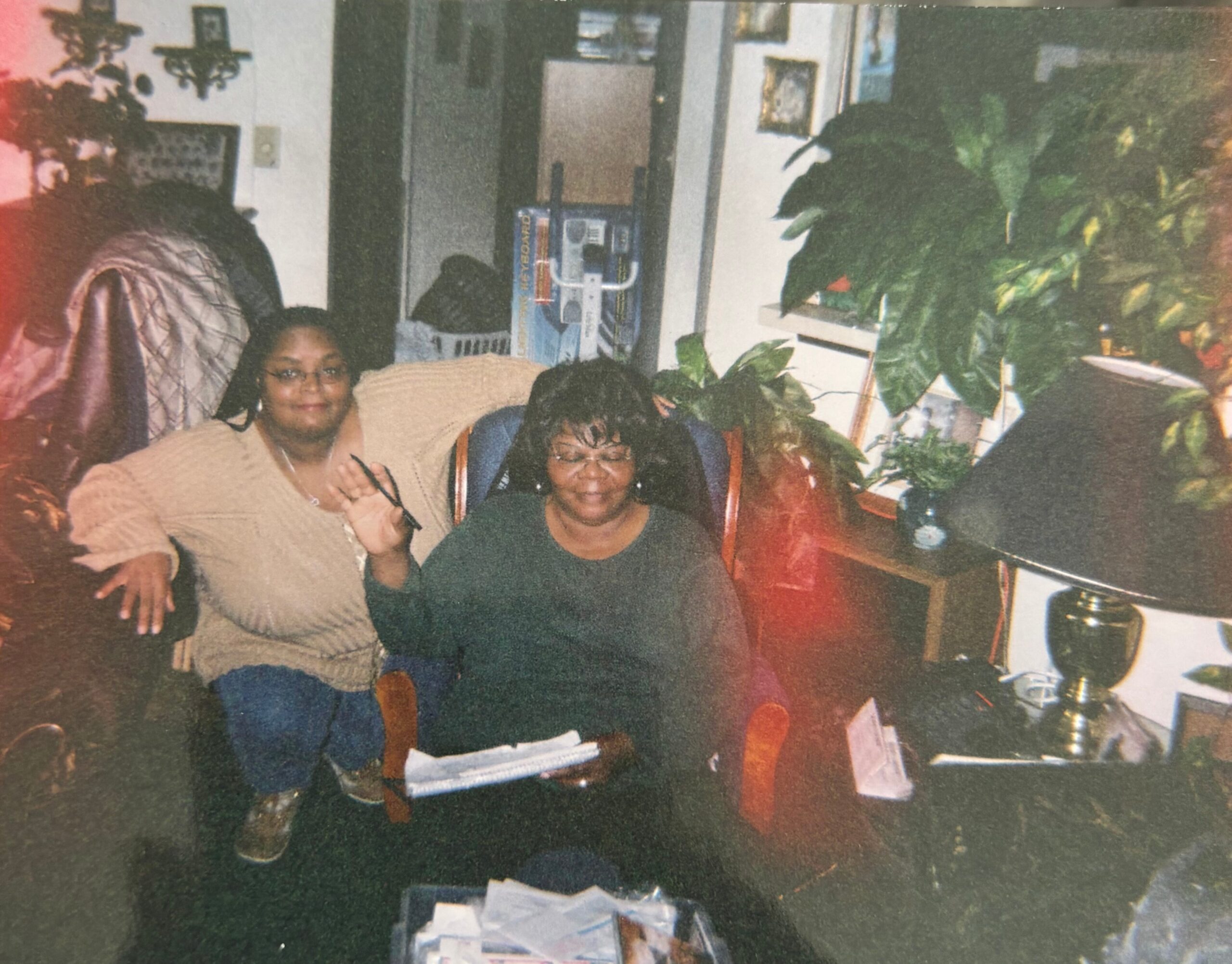 Two women sit on a beige couch in a plant-filled living room, smiling at the camera.