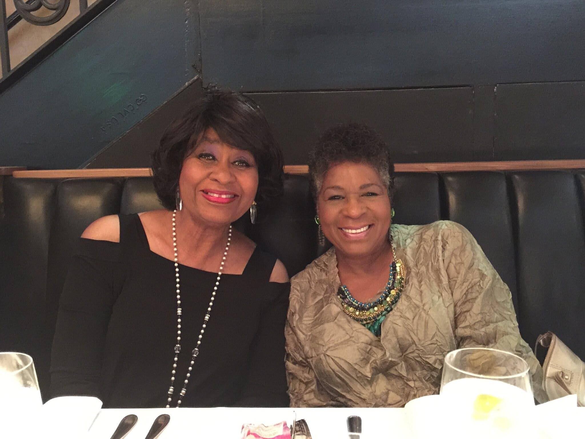 Two women smiling at a restaurant booth, wearing black and patterned outfits with beaded necklaces.