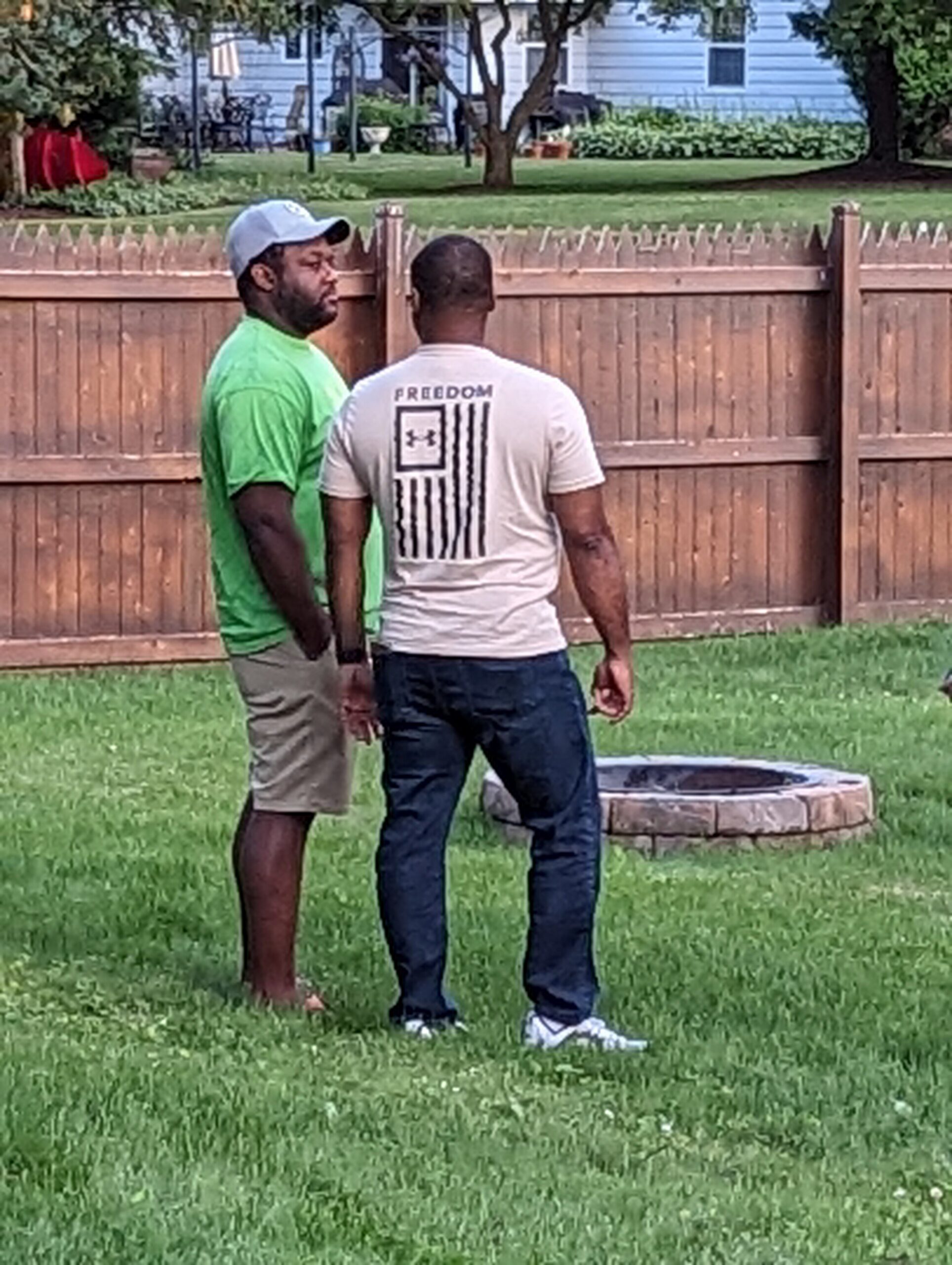 Two men stand facing each other on a green lawn near a wooden fence in a backyard; one in a bright green shirt and gray cap, the other in a white T-shirt with a 'FREEDOM' graphic on the back.