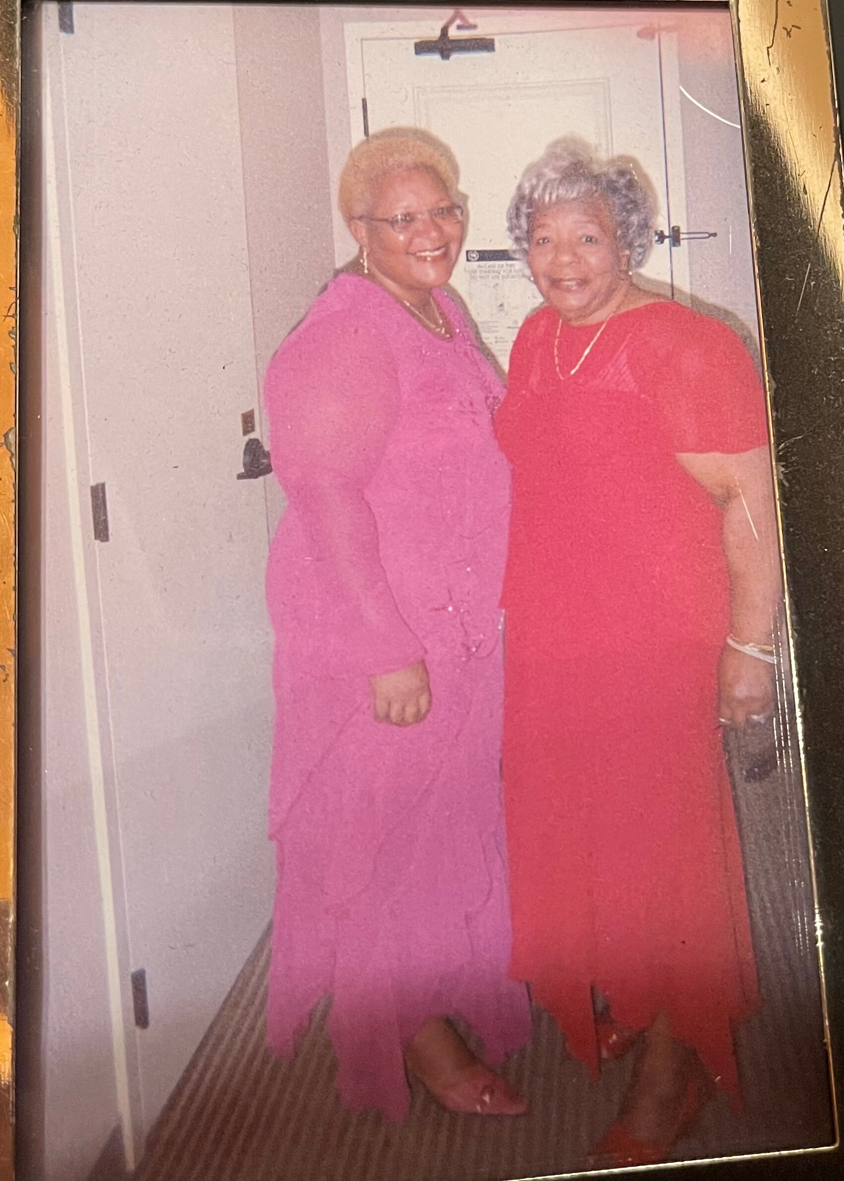 Two elderly women in bright dresses (pink and red) smiling together indoors in a home setting, posing by a door in a vintage photo.