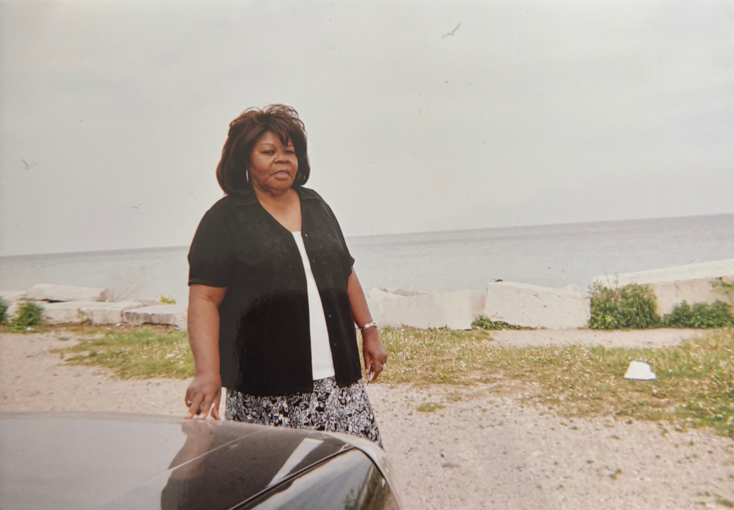 Woman standing beside a parked car near a rocky shoreline, with the sea in the background.