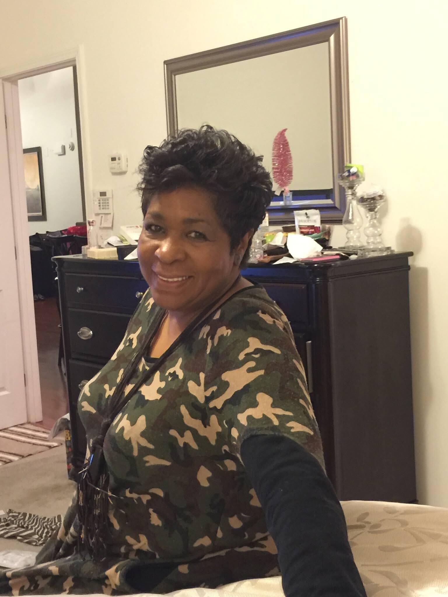 Smiling woman in camouflage top takes a selfie in a cluttered living room with a dresser and mirror behind her.