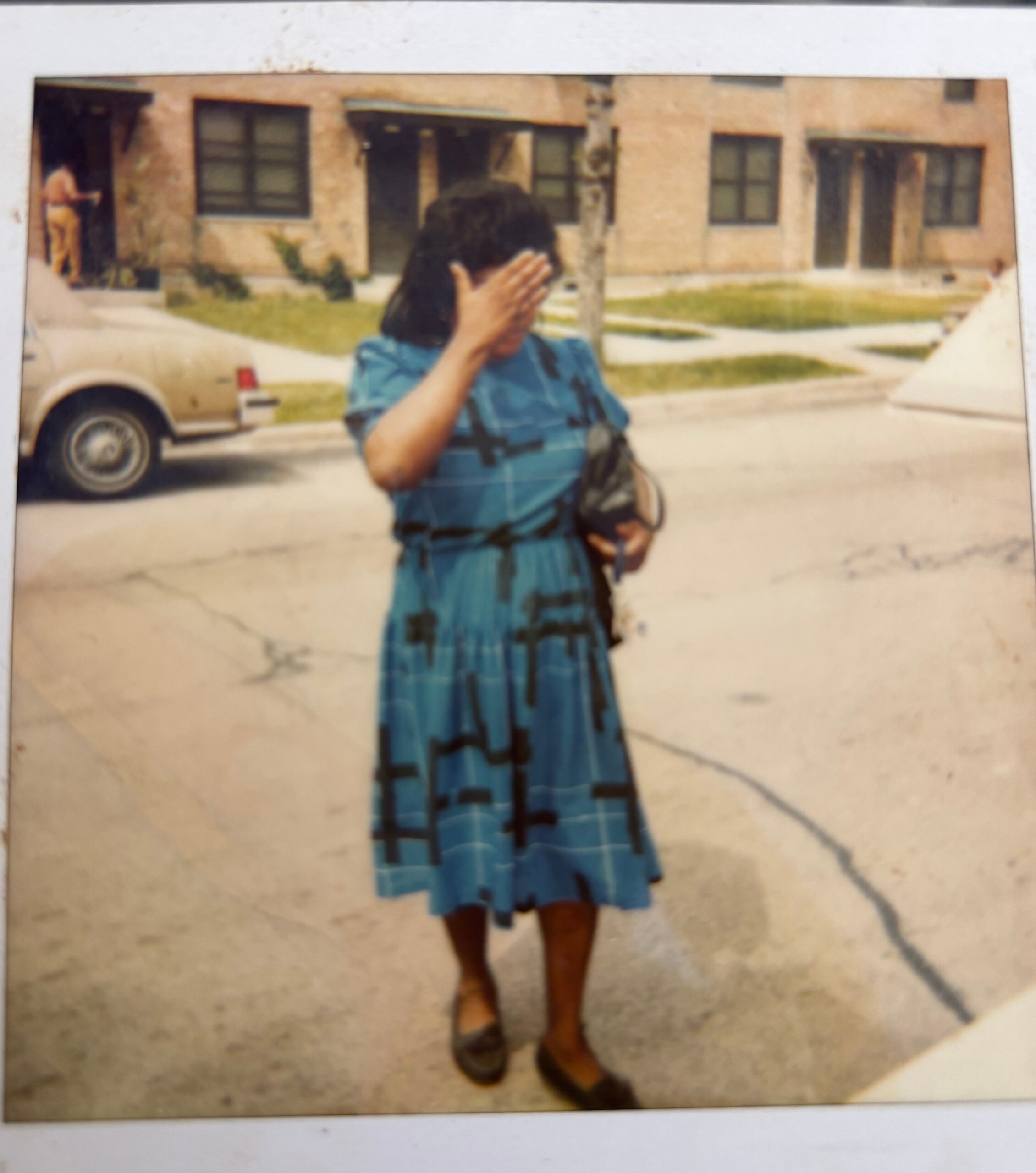 Woman in a blue patterned dress stands outside, hand raised to shield her face, with a beige building and parked car in the background.