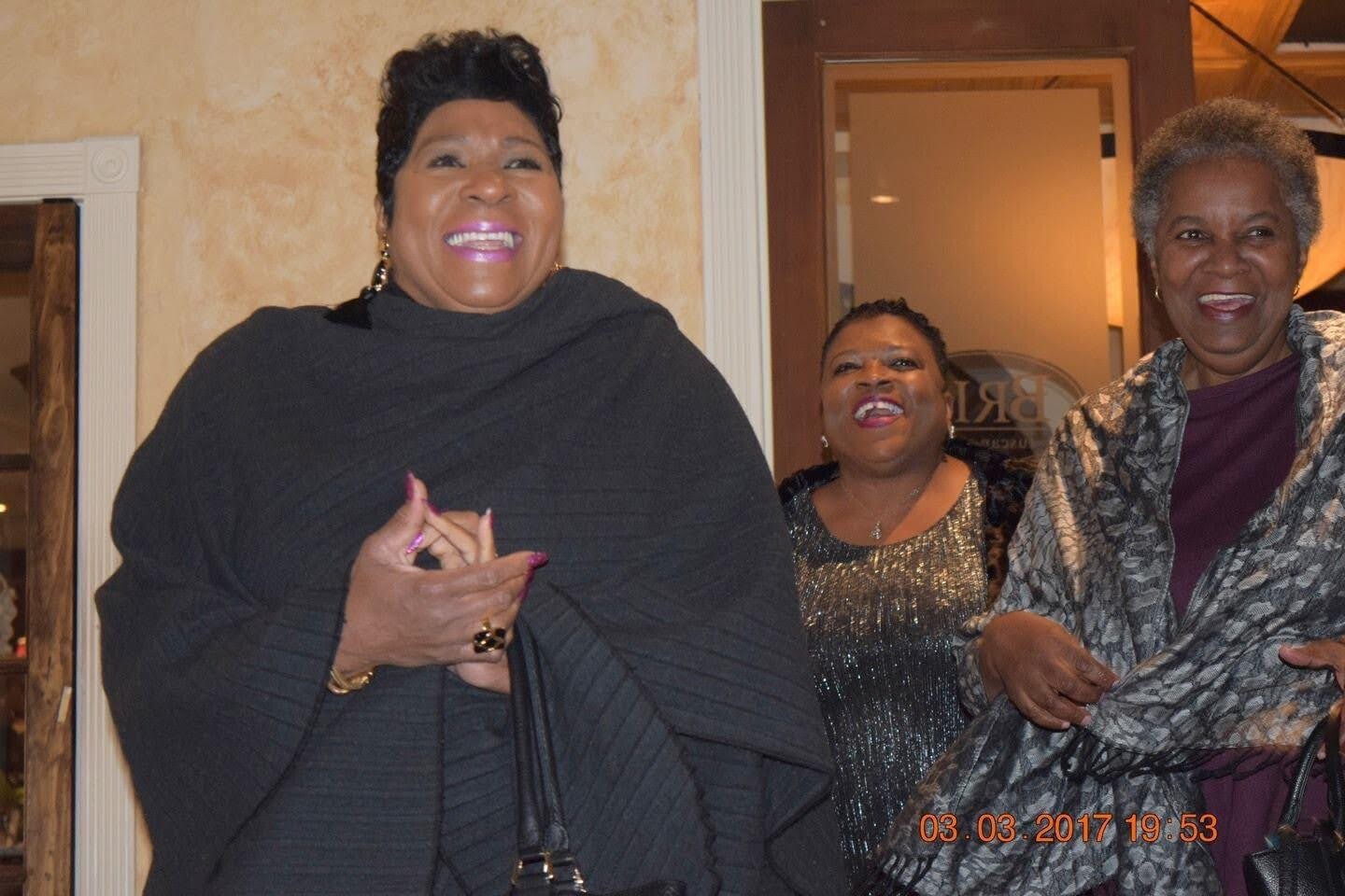 Three smiling Black women posing together indoors at a social event. Left: woman in a black poncho, center: woman in a sequined top, right: woman in a patterned shawl.