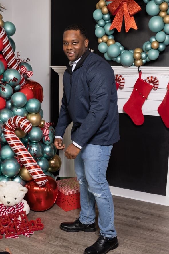Man in a navy pullover and jeans posing beside a Christmas display with teal and gold balloons, candy canes, stockings, and a wreath behind him.
