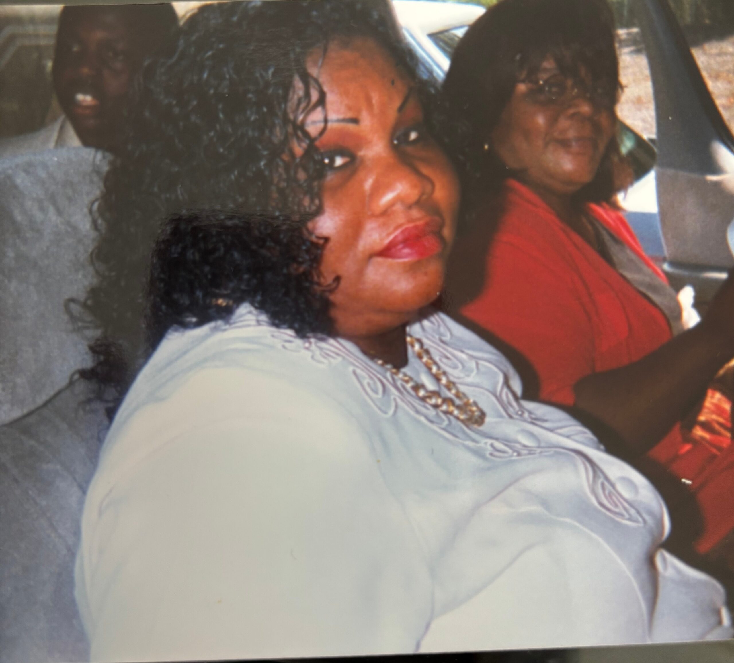 Two women sit in a car; the foreground woman has curly hair and red lipstick, the other wears a red top in the back seat.