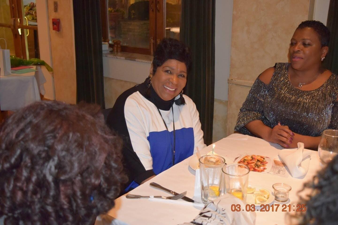 Two women sit at a white-tablecloth restaurant table, smiling, with plates and drinks in front of them.