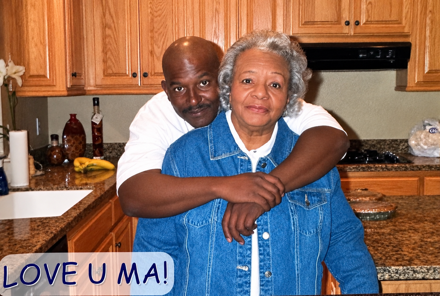 Man hugs an older woman from behind in a kitchen, both smiling at the camera. A warm, affectionate moment.
