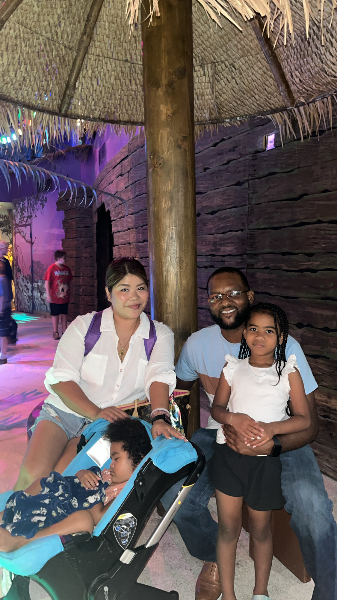 A family of three with a baby in a stroller posing for a photo under a thatched roof pillar in a tropical indoor setting.
