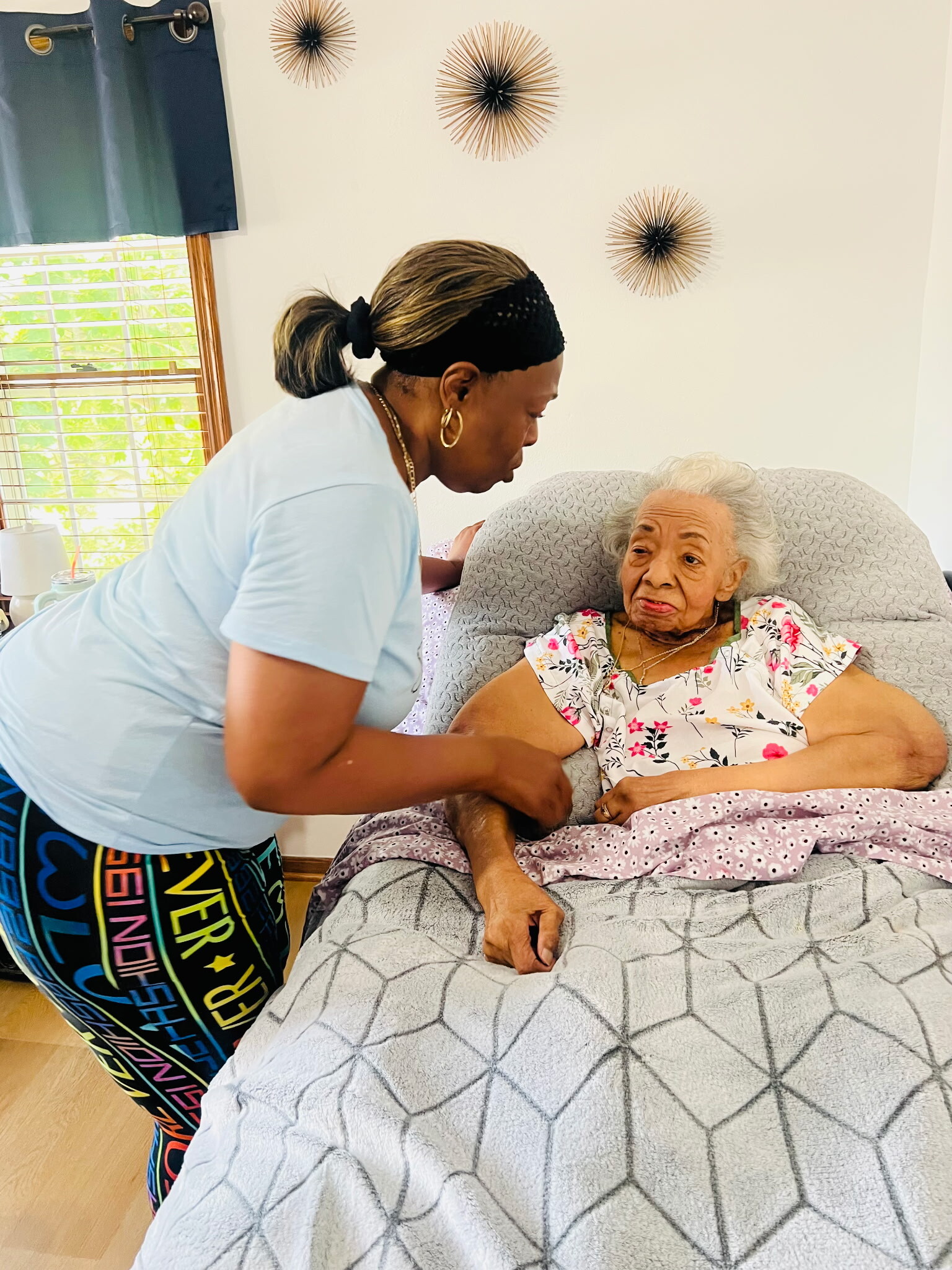 Caregiver leaning toward an elderly woman in bed, providing comfort in a sunlit room.