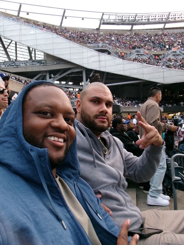 Two men sit in stadium seating at a sports event; one in a blue hoodie smiles at the camera, the other in a gray hoodie gestures with a peace sign in front of a packed crowd.