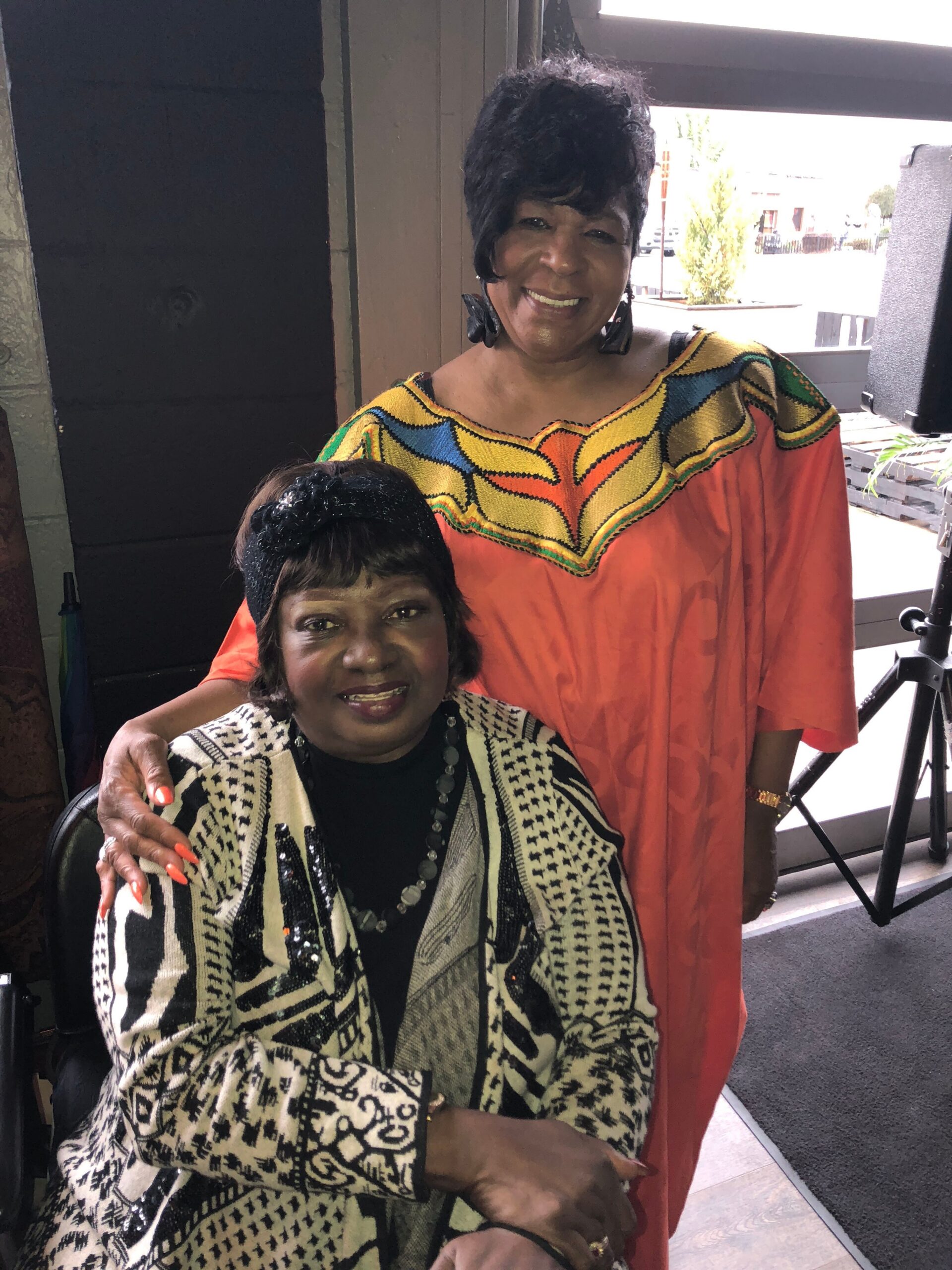 Two women posing indoors: a standing woman in a bright orange dashiki with a colorful neckline smiles beside a seated woman in a black‑and‑white patterned cardigan. A window and furniture are in the background.