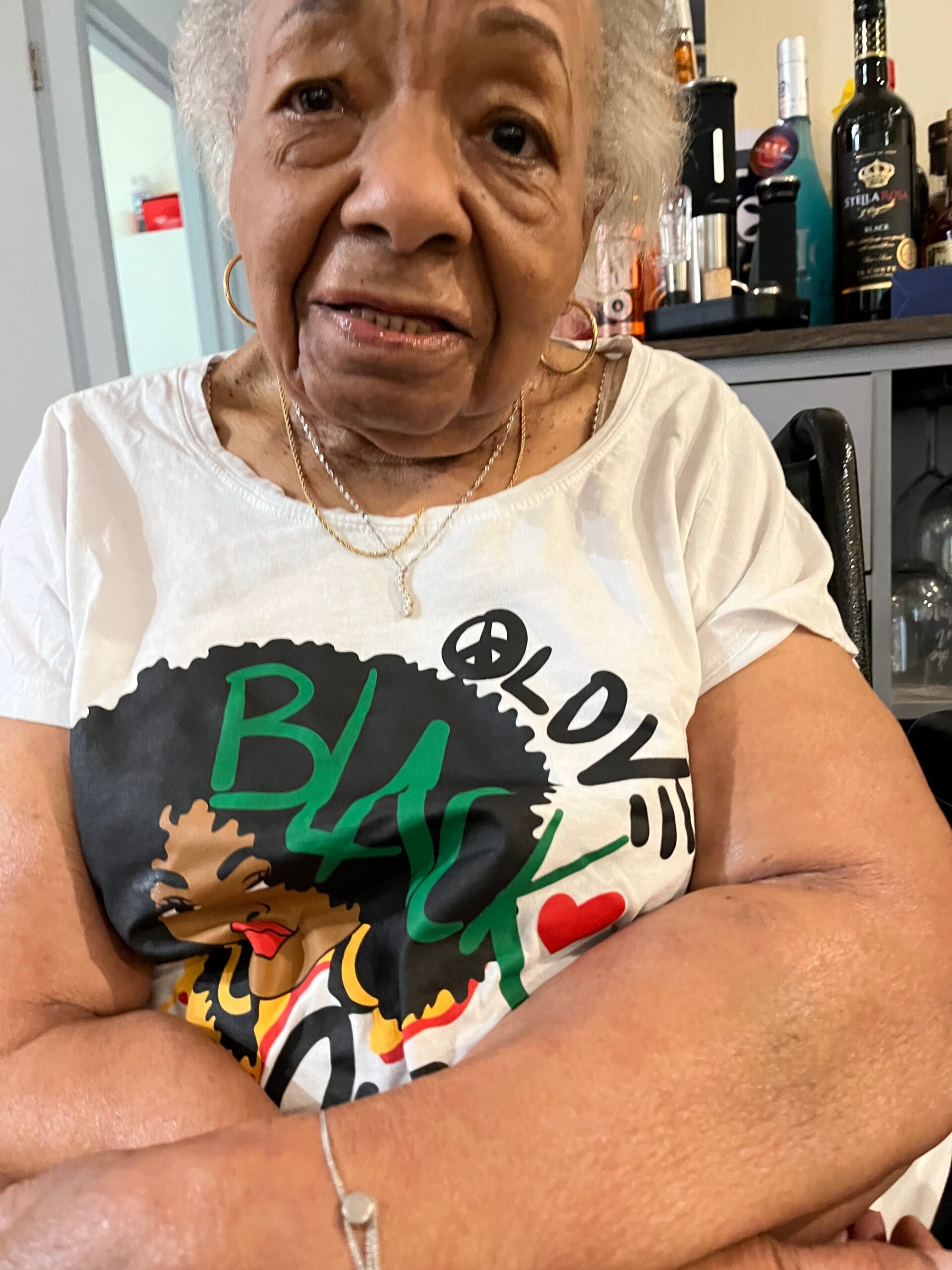 Close-up of an elderly woman with white hair wearing a white graphic tee, arms crossed, in a casual setting with bottles on a counter behind her