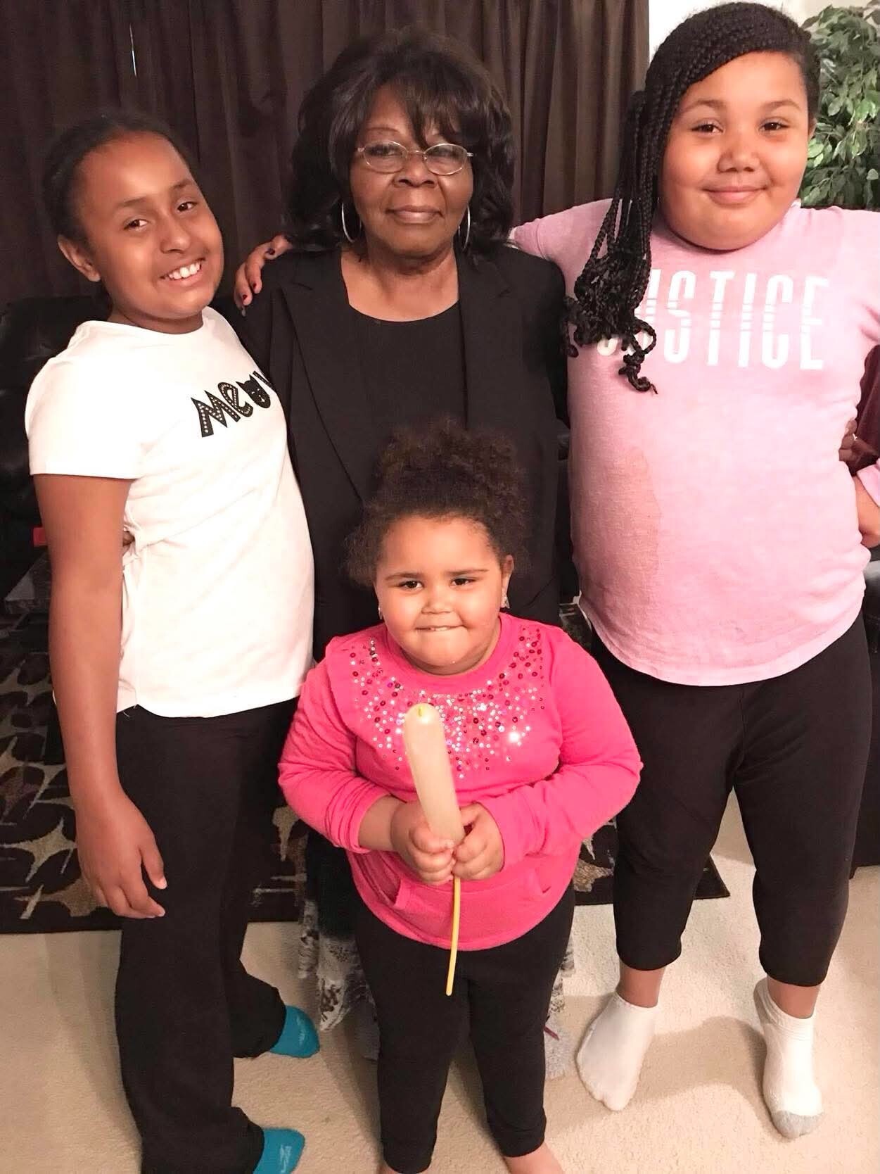 Grandmother in black with three smiling children indoors; the youngest holds a light-colored wand or baton in front.