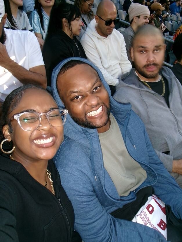 Group selfie at a sports stadium: smiling people seated together, foreground includes a woman with glasses and a man in a blue hoodie taking the photo.