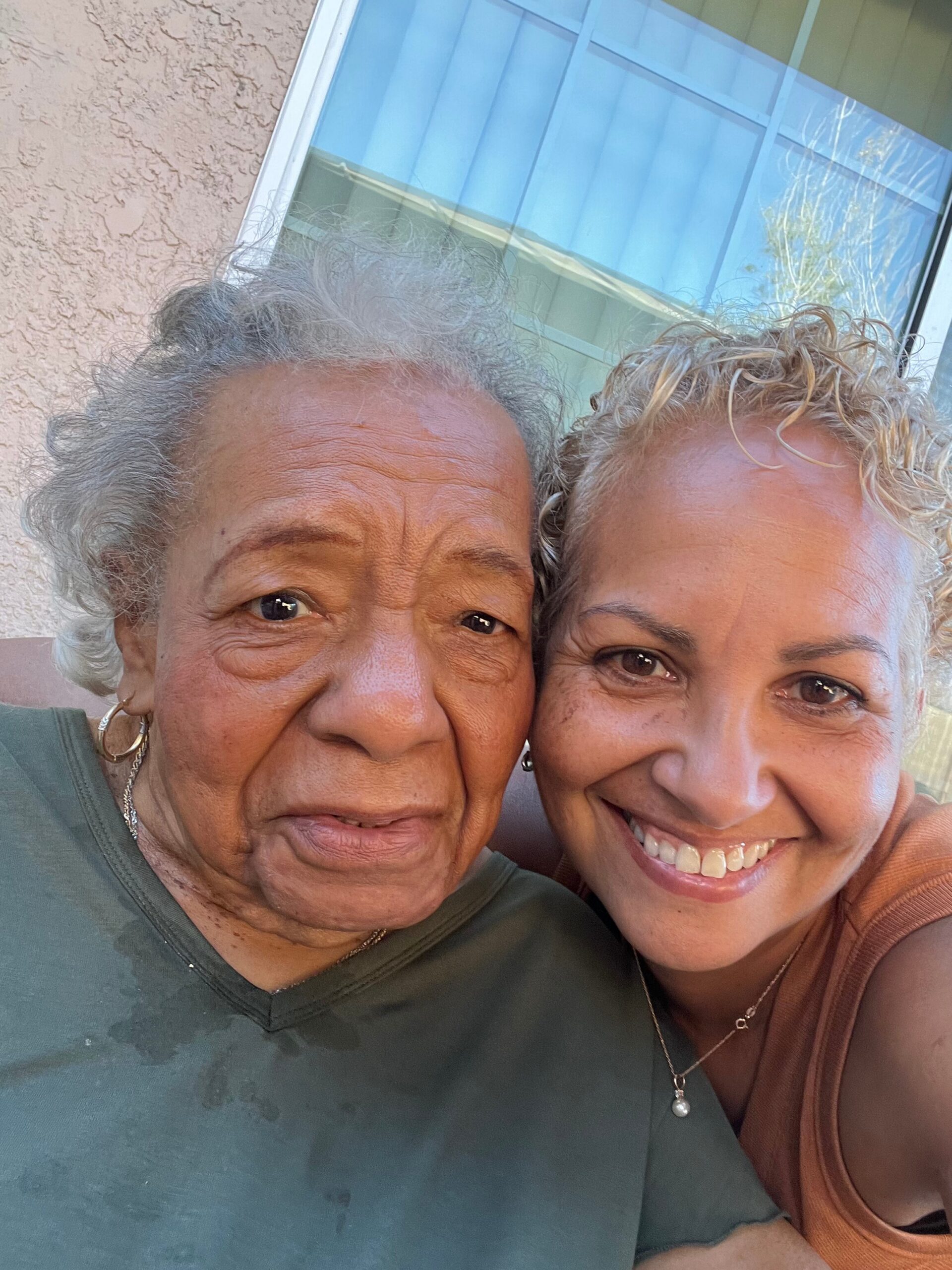 Two smiling women taking a close-up selfie outside, with a beige stucco wall and a blue-tinted window behind them behind them.