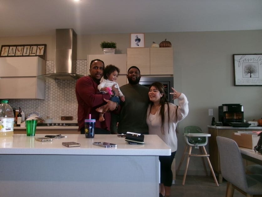 A family stands together in a modern kitchen, posing for the photo with a toddler held by one adult and another person making a peace sign next to them on the right.