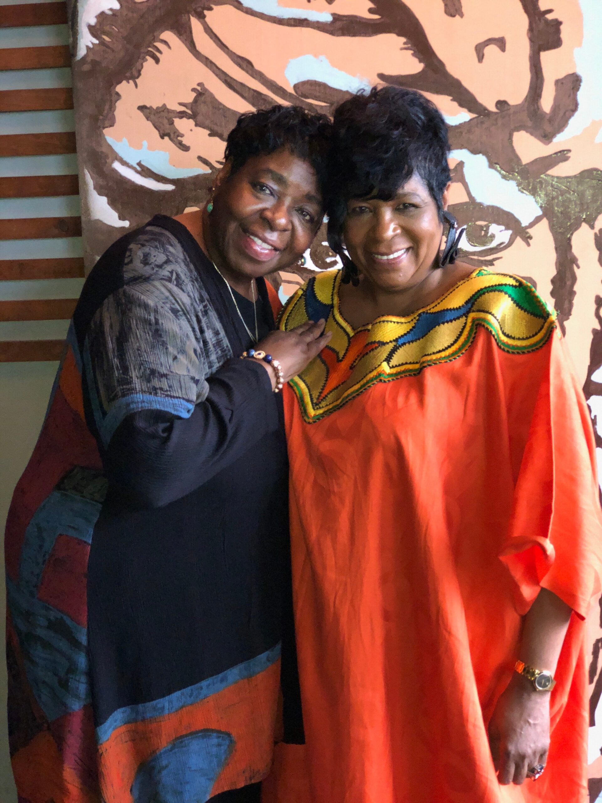 Two women posing and smiling together, one in a black outfit with a patterned shawl and the other in an orange dress with colorful embroidery, in front of a mural backdrop.