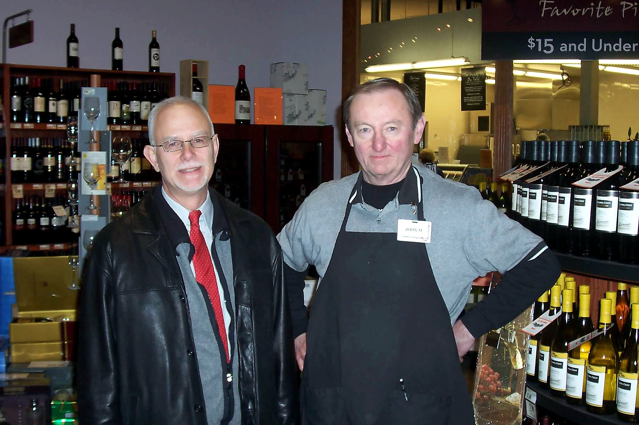 Two men pose behind a wine shop counter with bottles on shelves, one in a red tie and leather jacket, the other in a gray shirt and black apron.