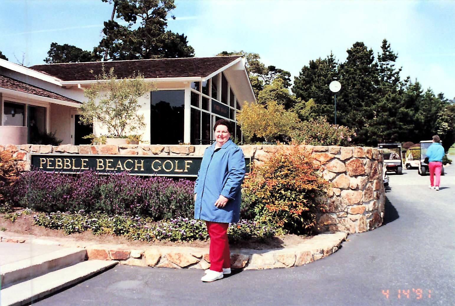 Woman in a blue coat and red pants stands beside a stone sign that reads Pebble Beach Golf, with landscaping behind him/her.