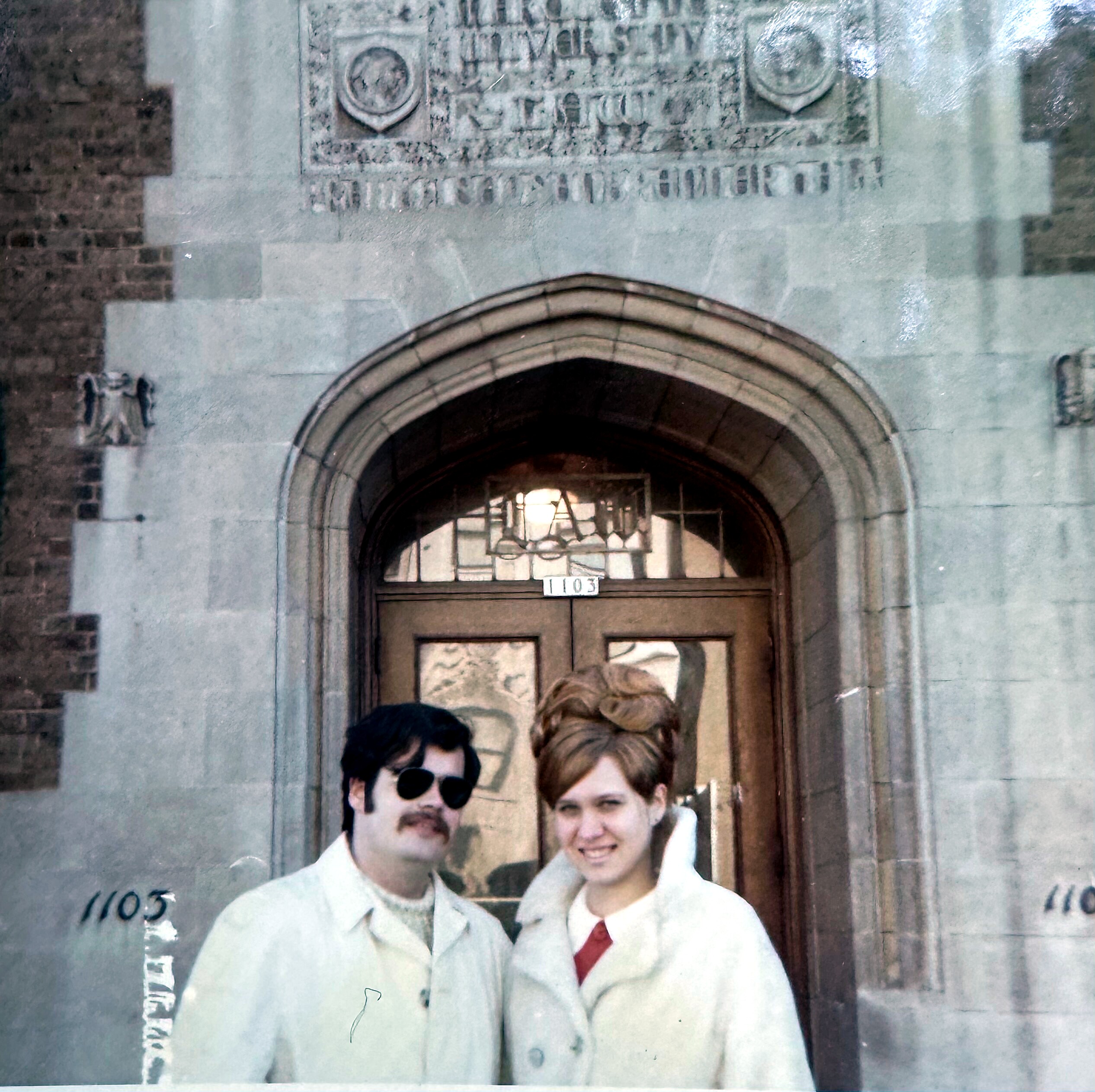 Two people pose by an arched doorway in a stone building, with a visible '1103' label nearby.