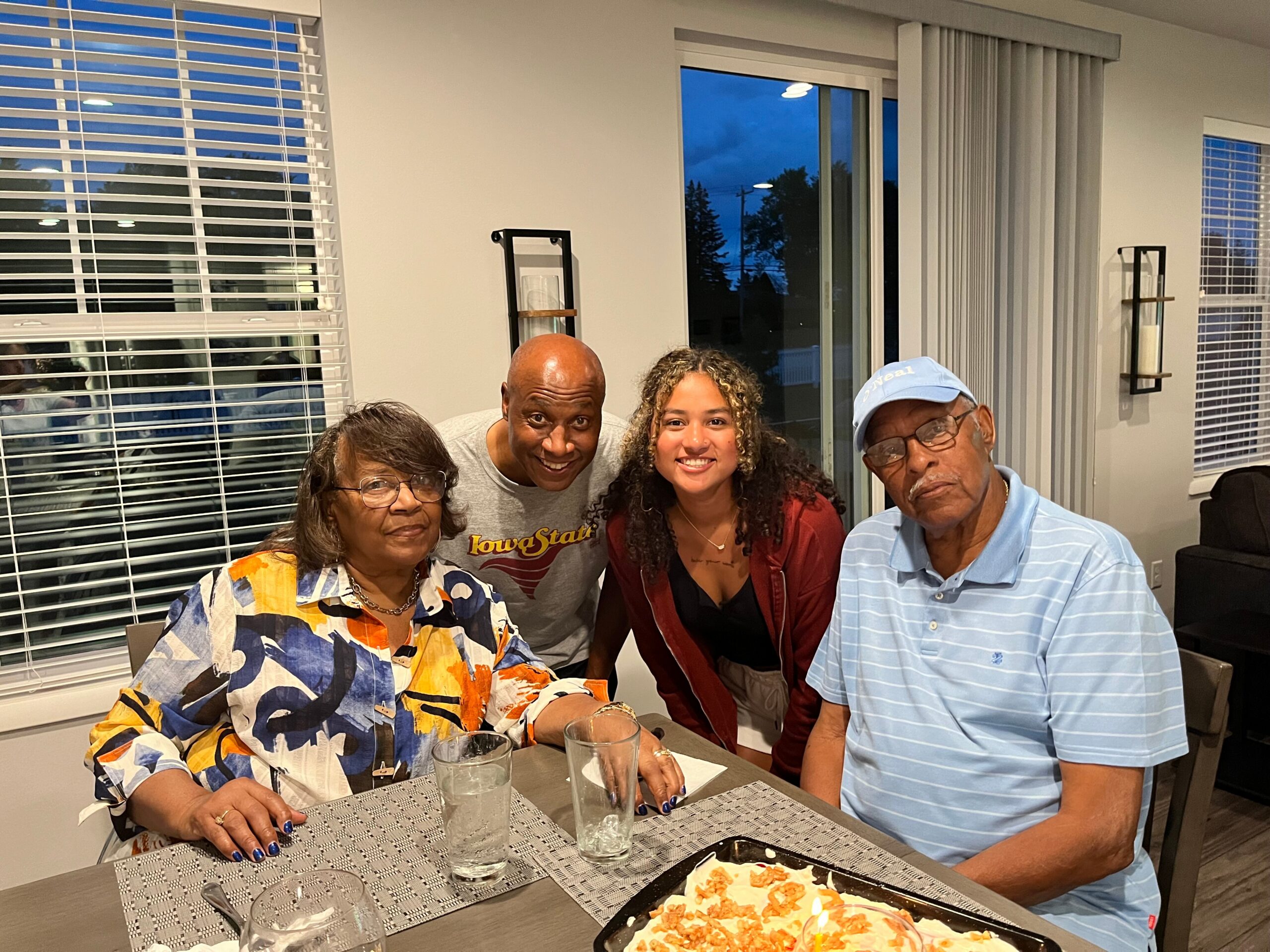Five adults sit around a dining table indoors, smiling for the camera; water glasses and plates with food are in the foreground are on the table.