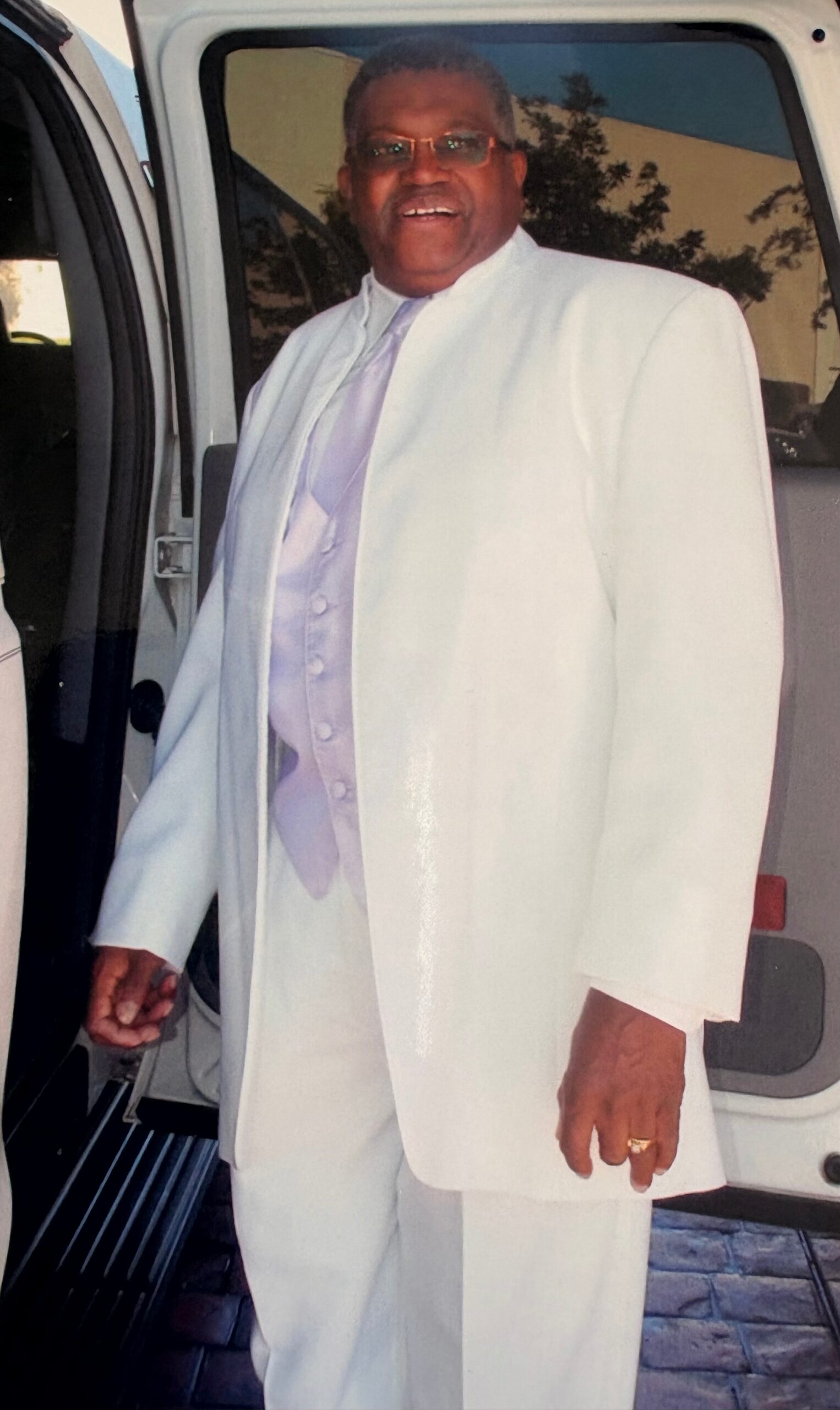 Man in a white suit with lavender vest and tie stands by an open car/van door, posing for the photo.