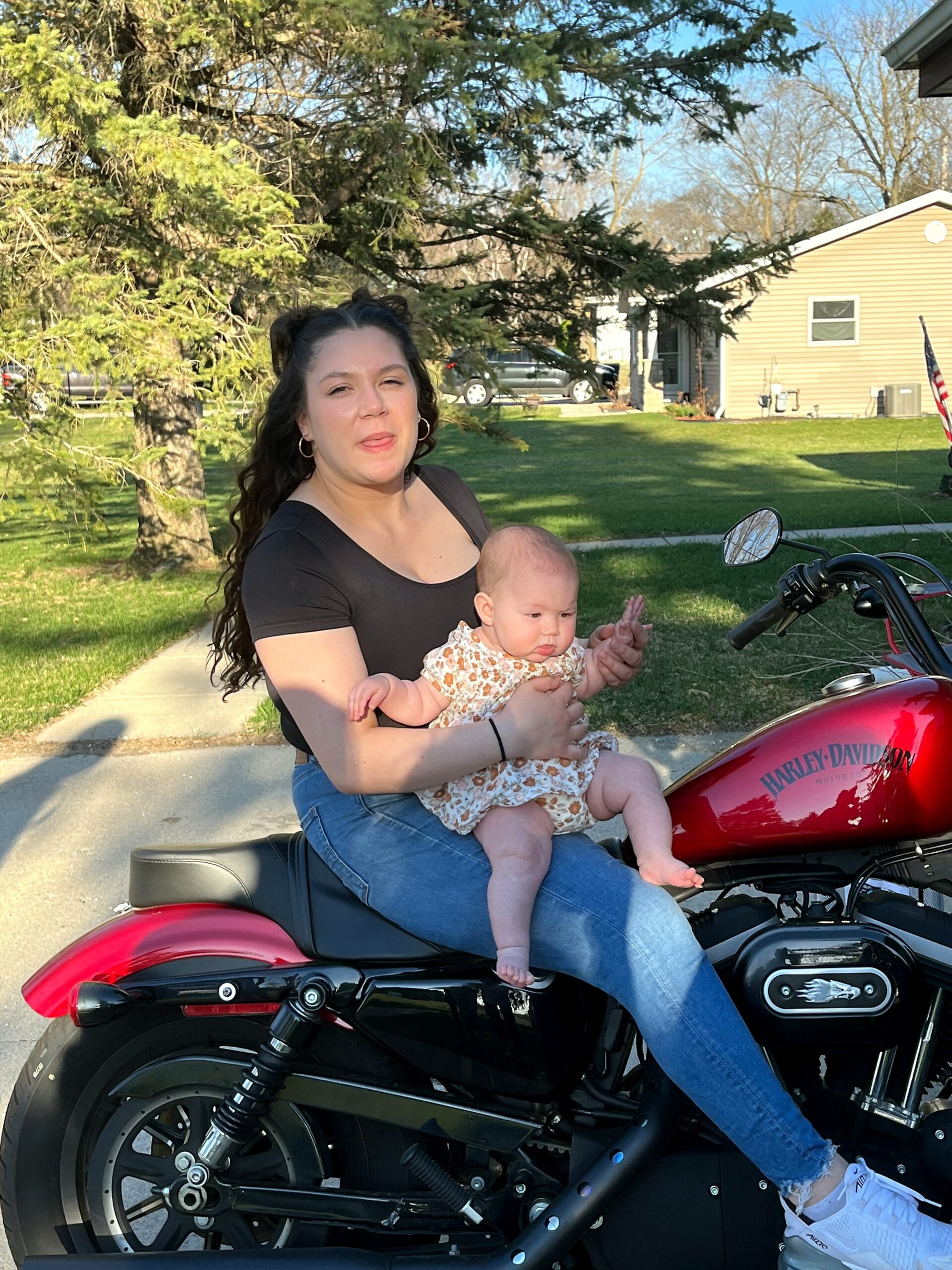 Woman holding a baby while sitting on a red Harley‑Davidson motorcycle in a suburban driveway on a sunny day.