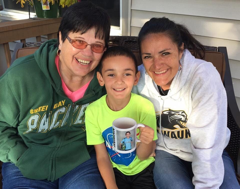Three family members—the grandmother, mother, and young son—sit on a porch smiling at the camera; the boy holds a mug with a photo on it.