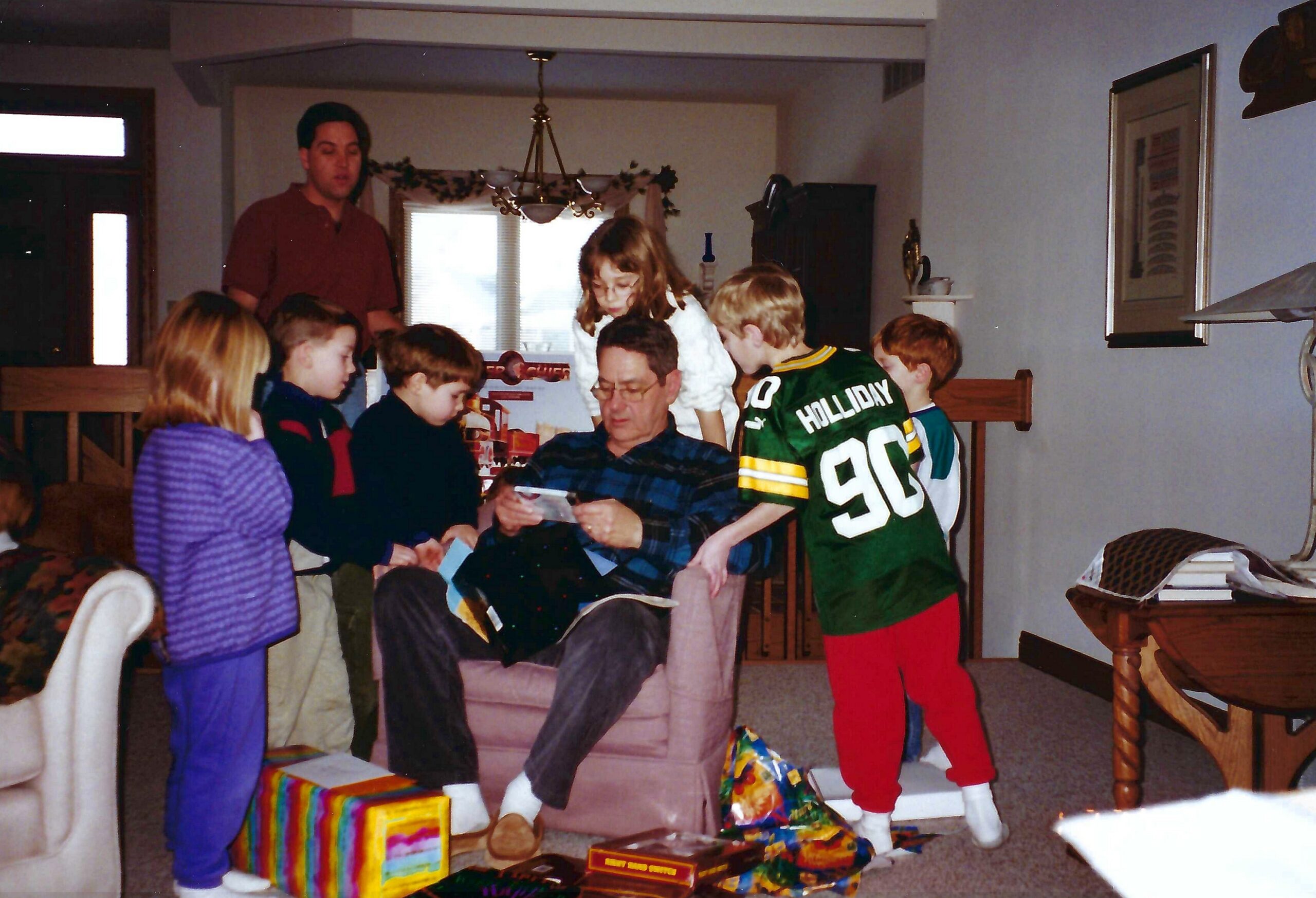 Man sits in a chair while children hand him wrapped gifts at a family holiday gathering in a living room