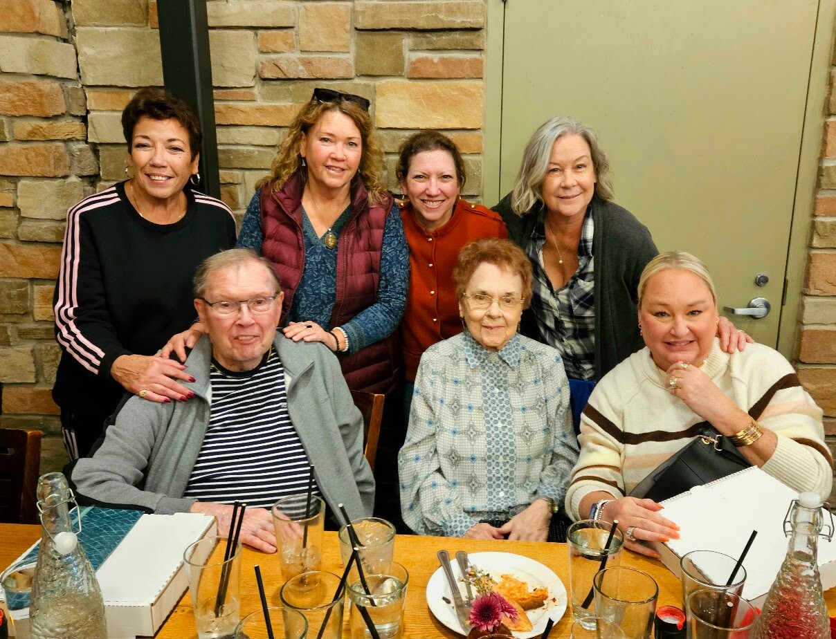 Seven adults smiling at a restaurant table, posing for a group photo with drinks and plates around them on a brick-walled background.