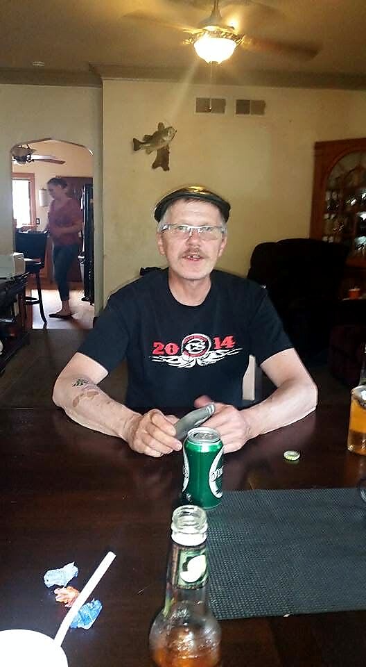 Man wearing a cap and glasses sits at a wooden table with a green beer can in front, holding an item, in a living room with another person visible in a doorway.