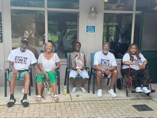 Five people of varying ages and backgrounds sit on a sidewalk bench outside a building, smiling at the camera.