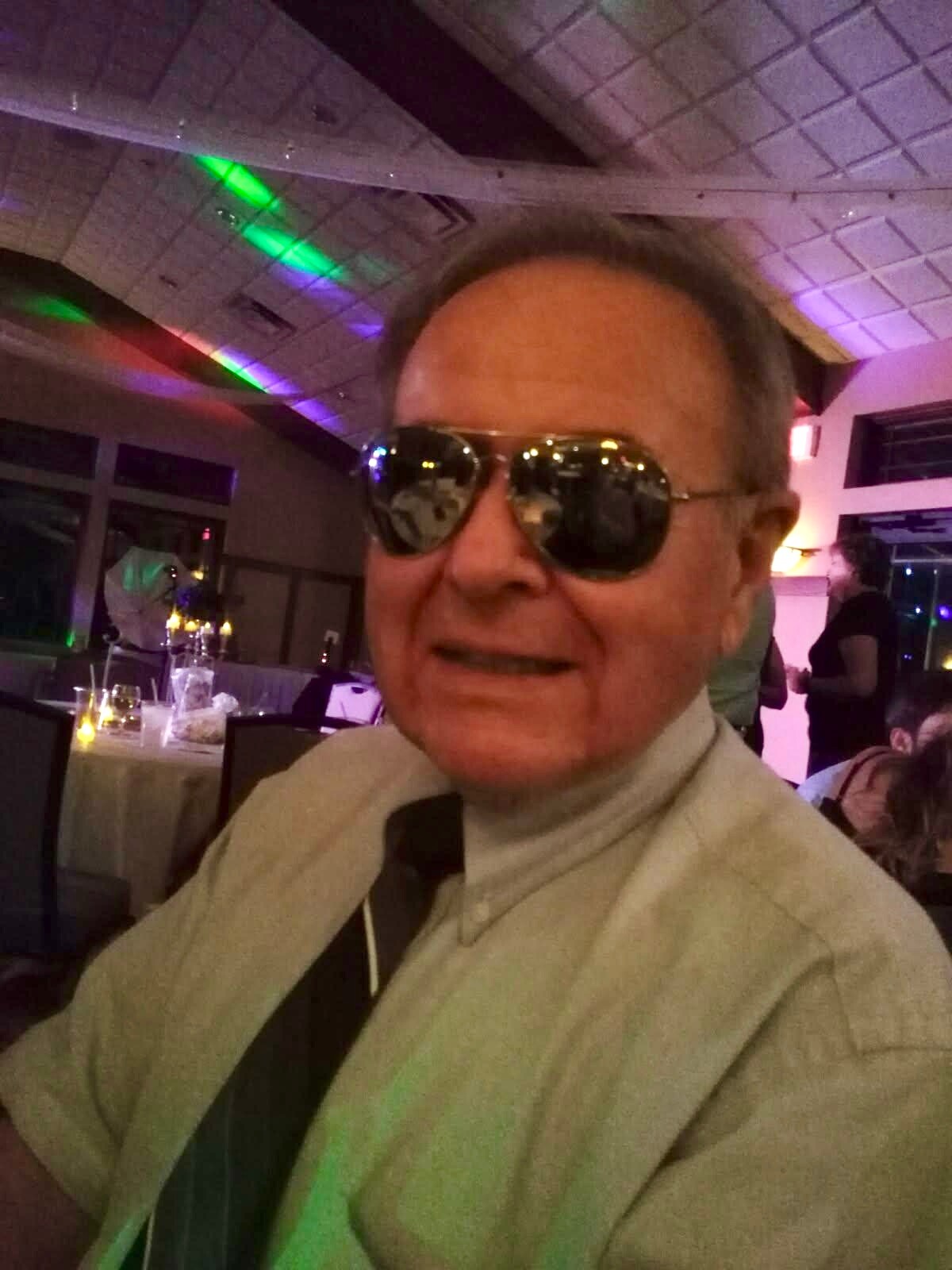 Smiling man in a beige suit and sunglasses at a dimly lit party with colorful lights in the background.