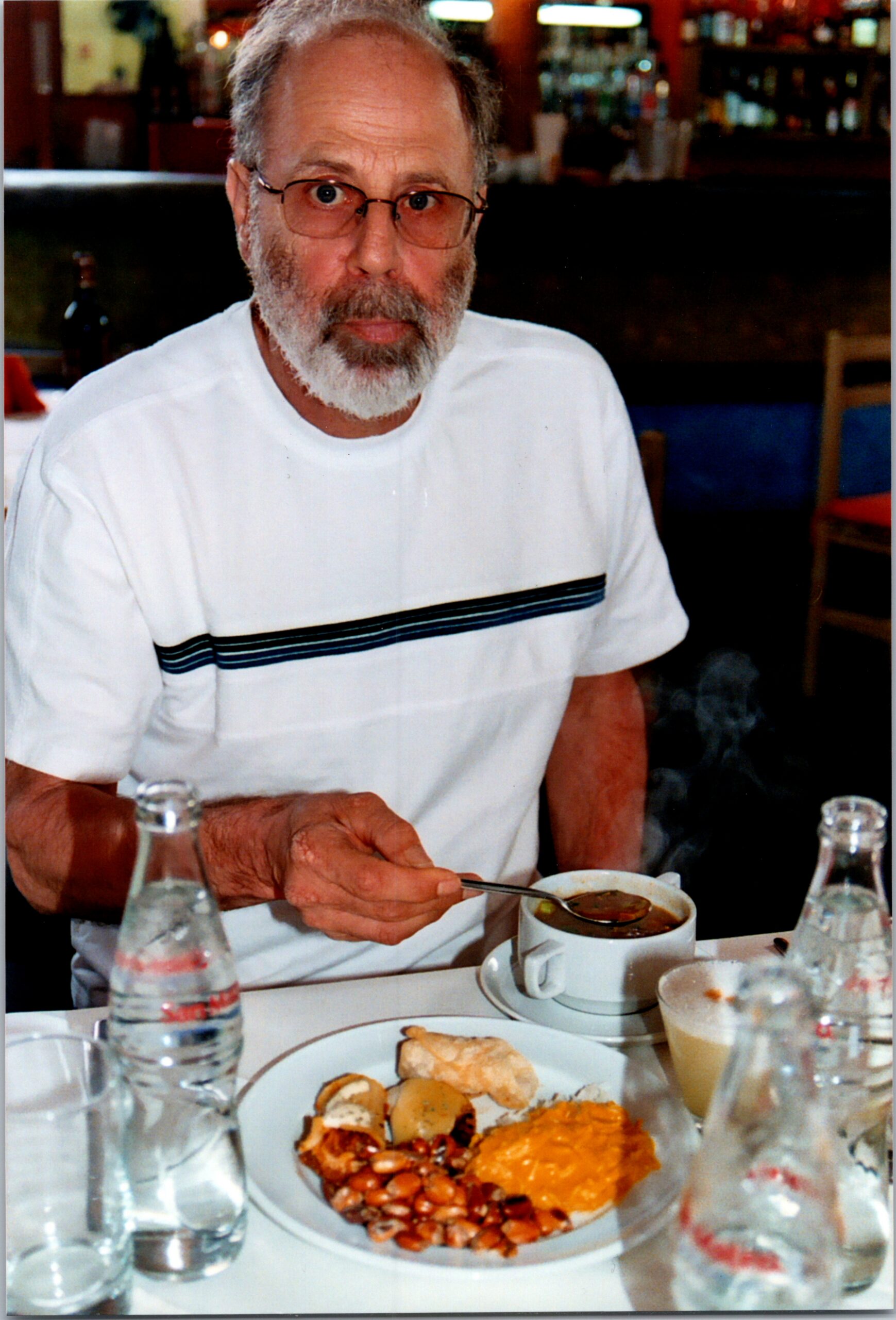 Older man with glasses and a white beard sits at a restaurant table, spooning soup from a bowl with a plate of beans and vegetables nearby.