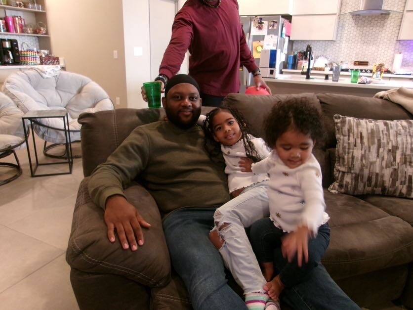 Father relaxing on a brown couch with his two young daughters in a modern living room and open kitchen in the background.