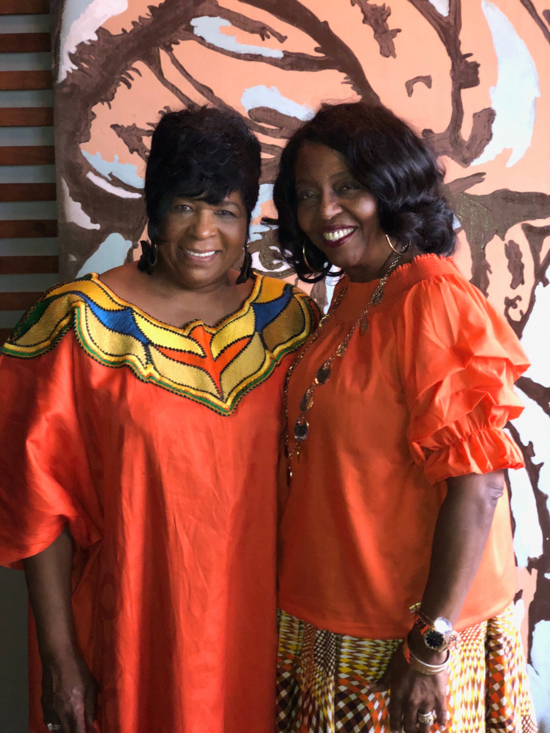 Two women in bright orange outfits smile for the camera, standing in front of a patterned mural; one wears a colorful dash-pattern skirt visible at the bottom.